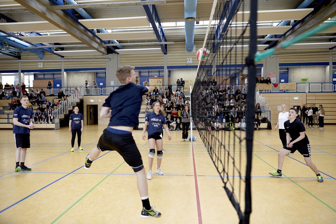 Hallen-Volleyball: Spieler springt zum Spike über Netz, Gegner in Abwehrstellung, Zuschauer auf Tribüne