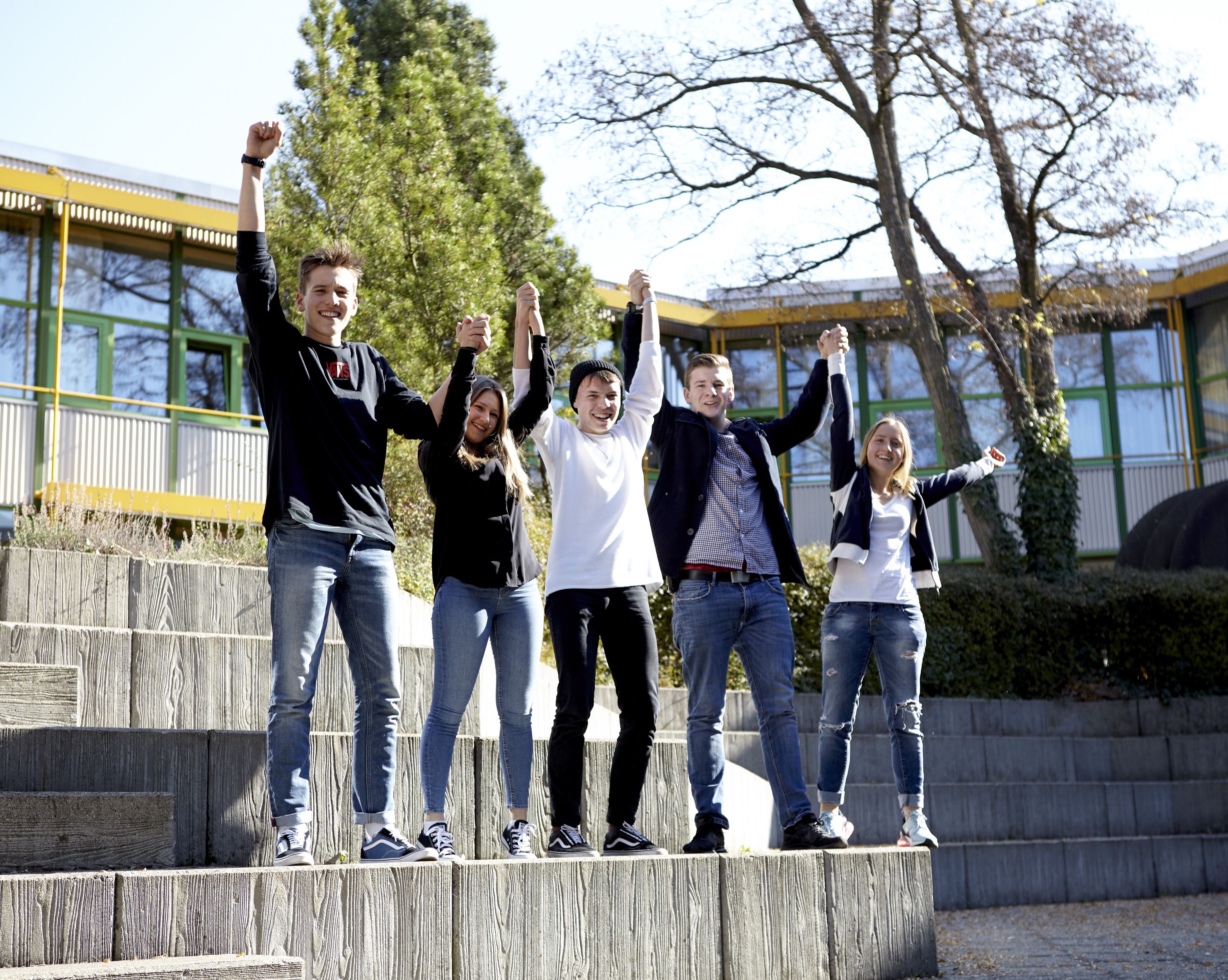 Fünf Jugendliche stehen auf Treppenstufen vor einem Schulgebäude, halten sich an den Händen und jubeln
