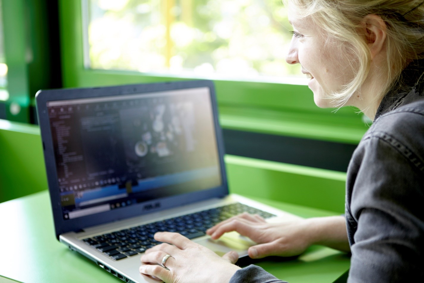 Lächelnde junge Frau tippt am Laptop an grünem Tisch vor einem Fenster.