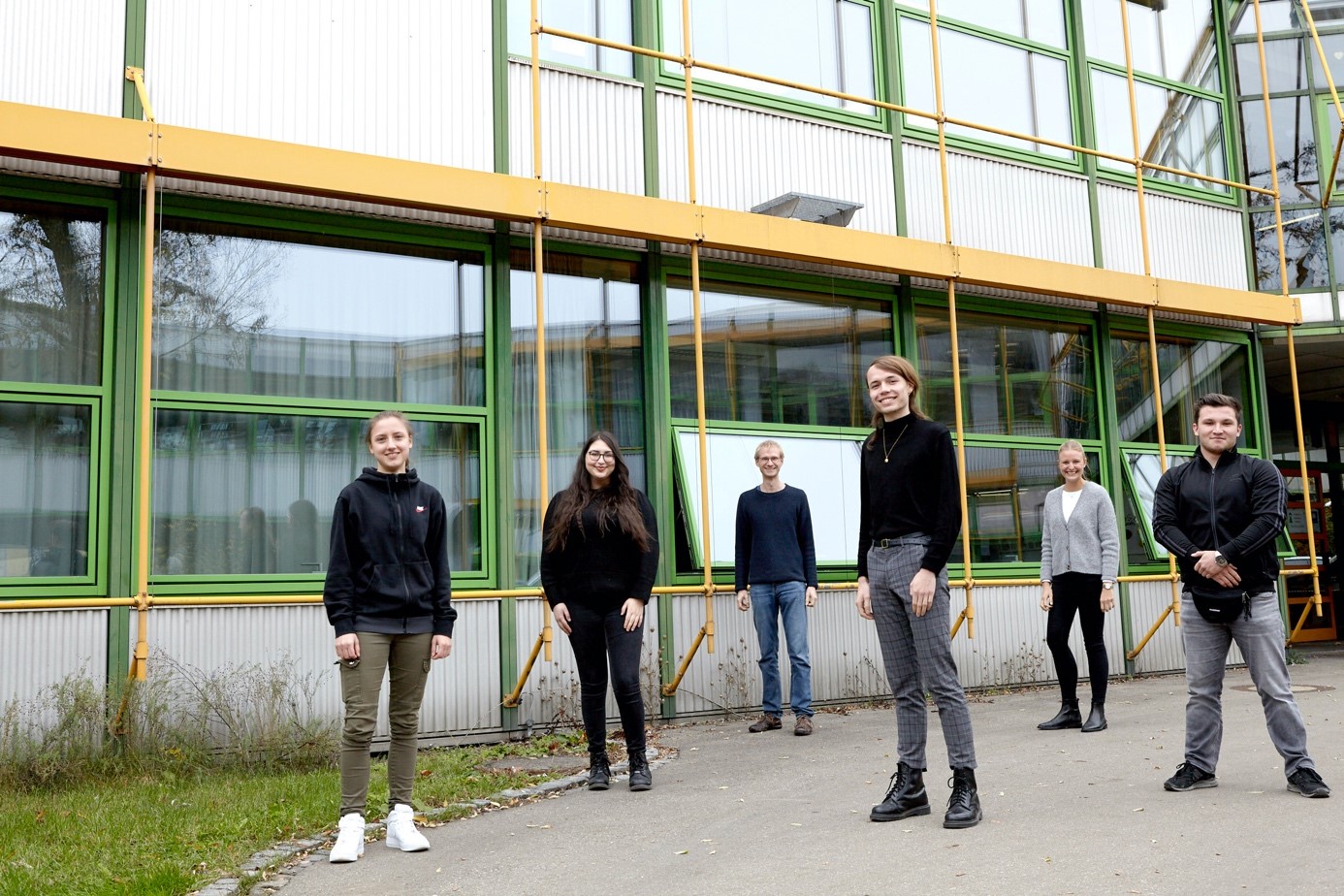 Sechs Personen stehen mit Abstand vor einem Gebäude mit grün-gelben Fensterrahmen, lächelnd