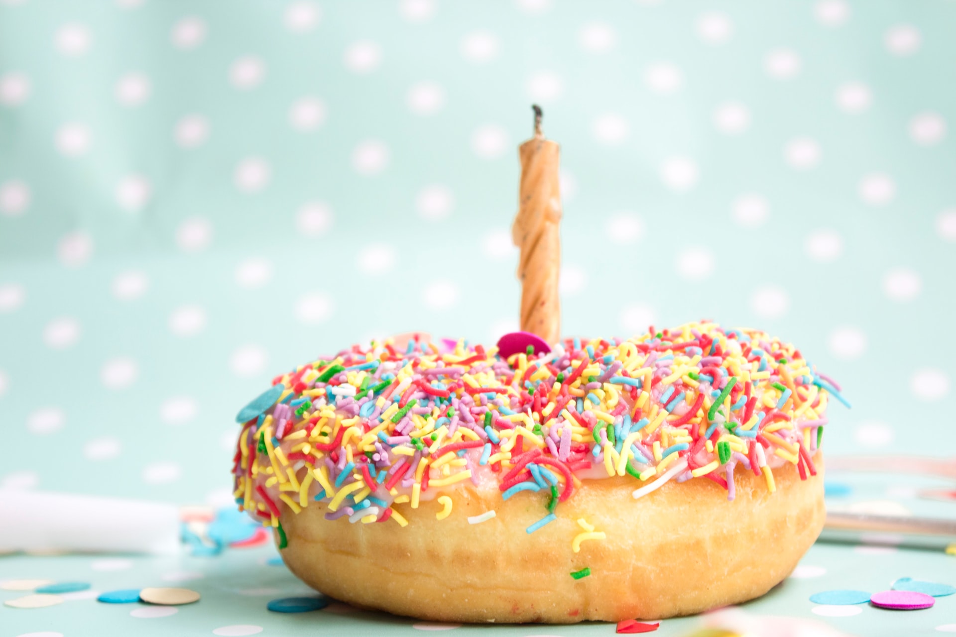 Donut mit rosa Glasur und bunten Streuseln, abgebrannte Kerze steckt in der Mitte vor pastellblauem, gepunktetem Hintergrund