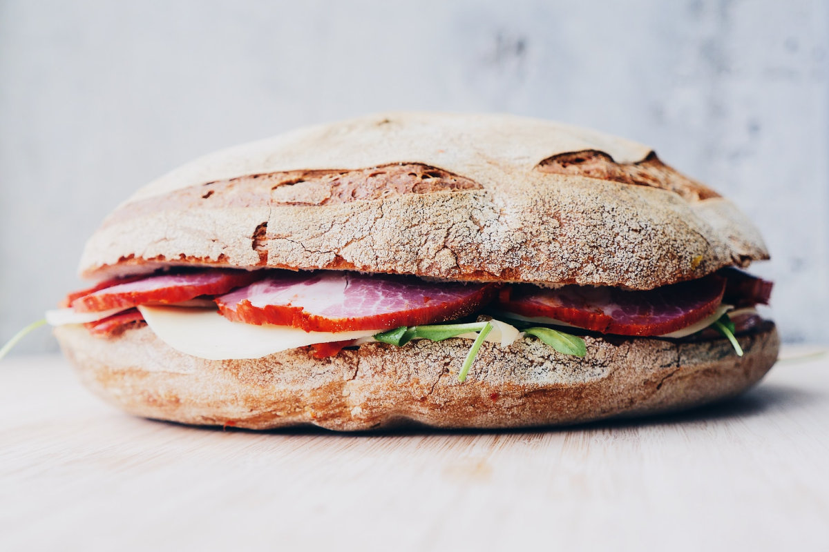 Rustikales Ciabatta-Sandwich mit rotem Aufschnitt, Käsescheiben und Rucola auf hellem Holzbrett.