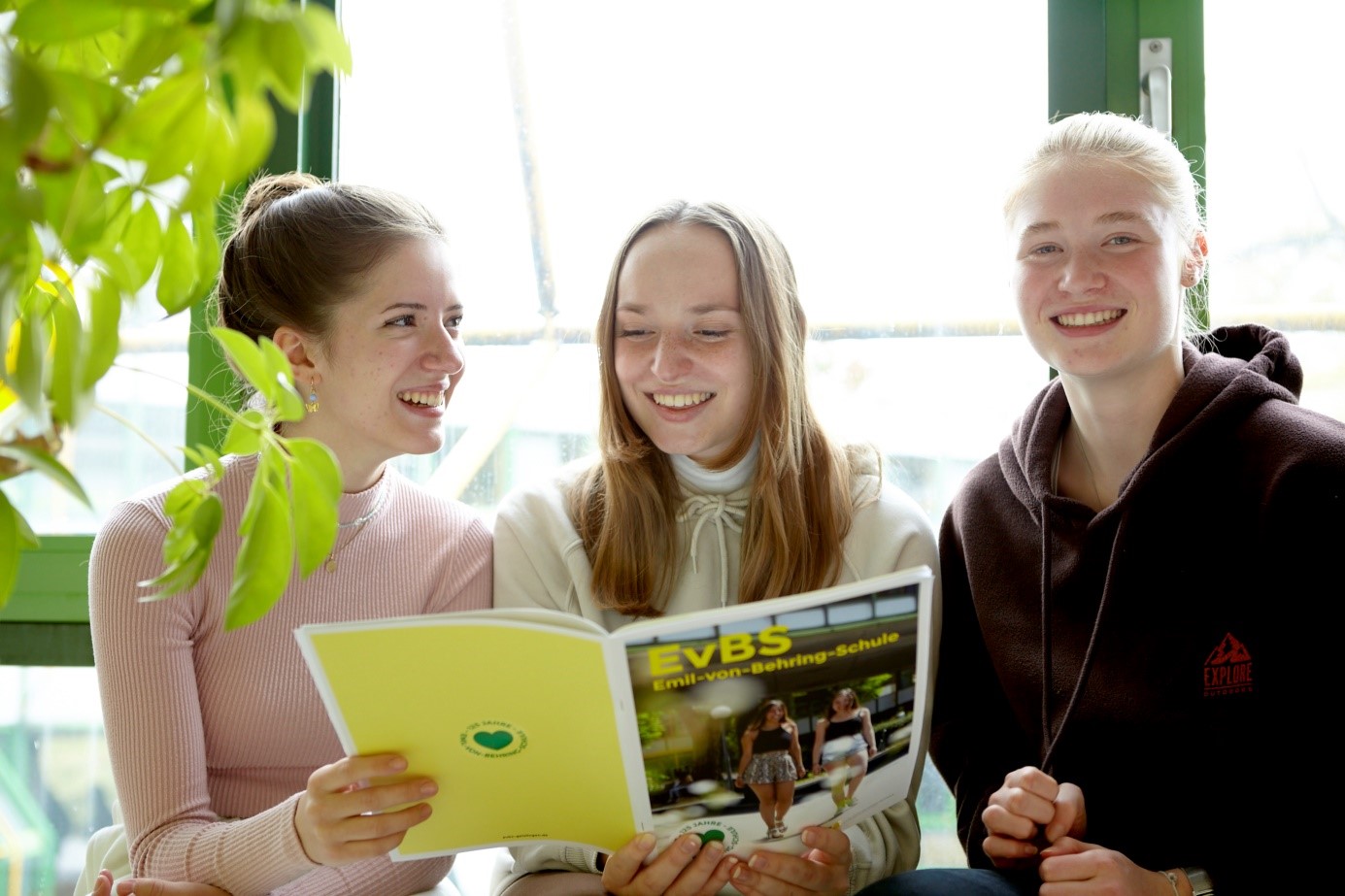 Drei lächelnde Schülerinnen sitzen am Fenster und lesen gemeinsam eine gelbe Schulbroschüre (EVBS).