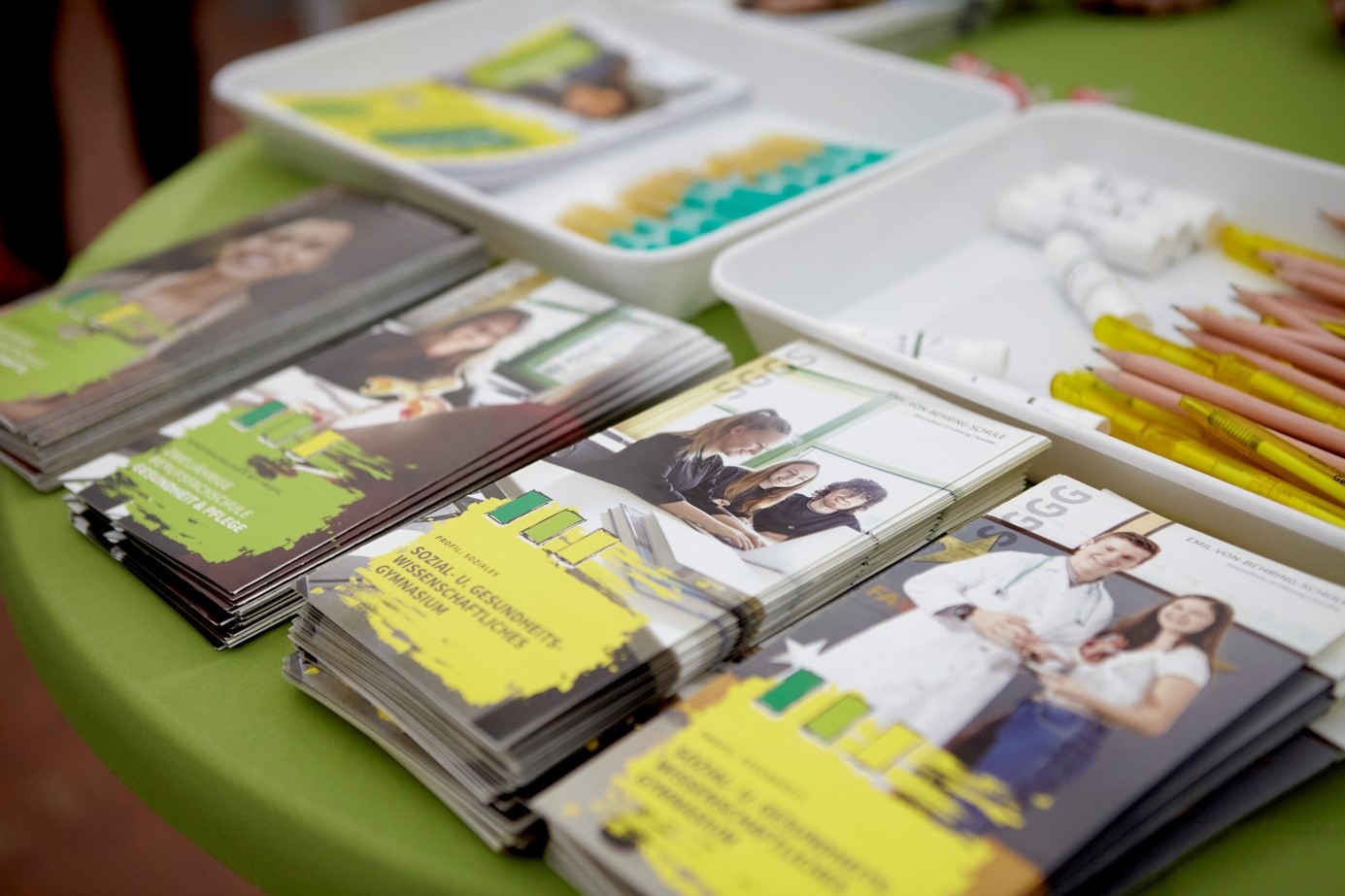 Gestapelte Flyer mit Schüler- und Arztporträts auf grünem Tisch, daneben gelbe Kugelschreiber und Tabletts