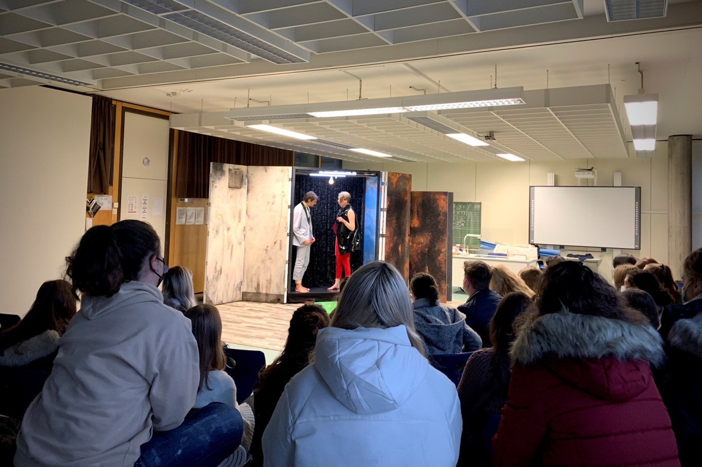 Schüler im Klassenraum verfolgen auf kleiner Bühne ein Gespräch zwischen zwei Darstellern.