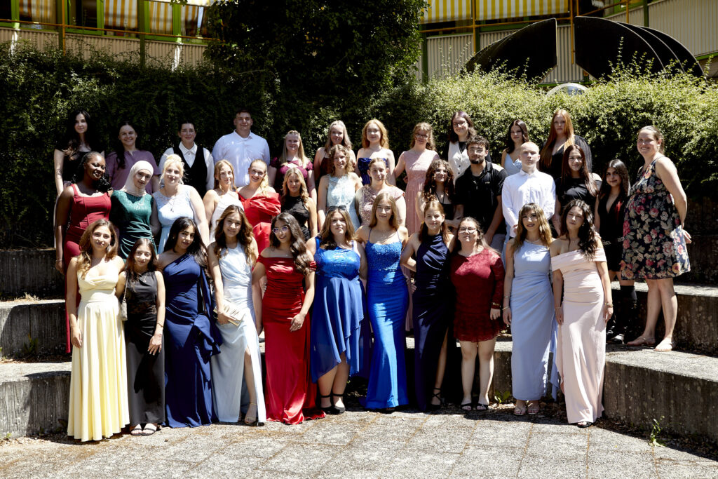 Eine große Gruppe von Absolventinnen und Absolventen steht in eleganten Kleidern und Anzügen auf einer Außentreppe der Schule. Sie lächeln bei sonnigem Wetter in die Kamera.