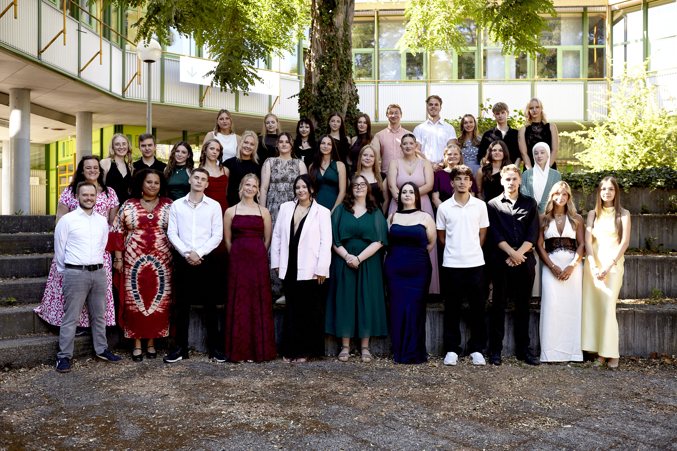 Ein Jahrgang von Schülerinnen und Schülern posiert für ein Abschlussfoto auf den Steinstufen des Schulhofs. Alle tragen schicke Abendgarderobe wie Anzüge und lange Kleider.