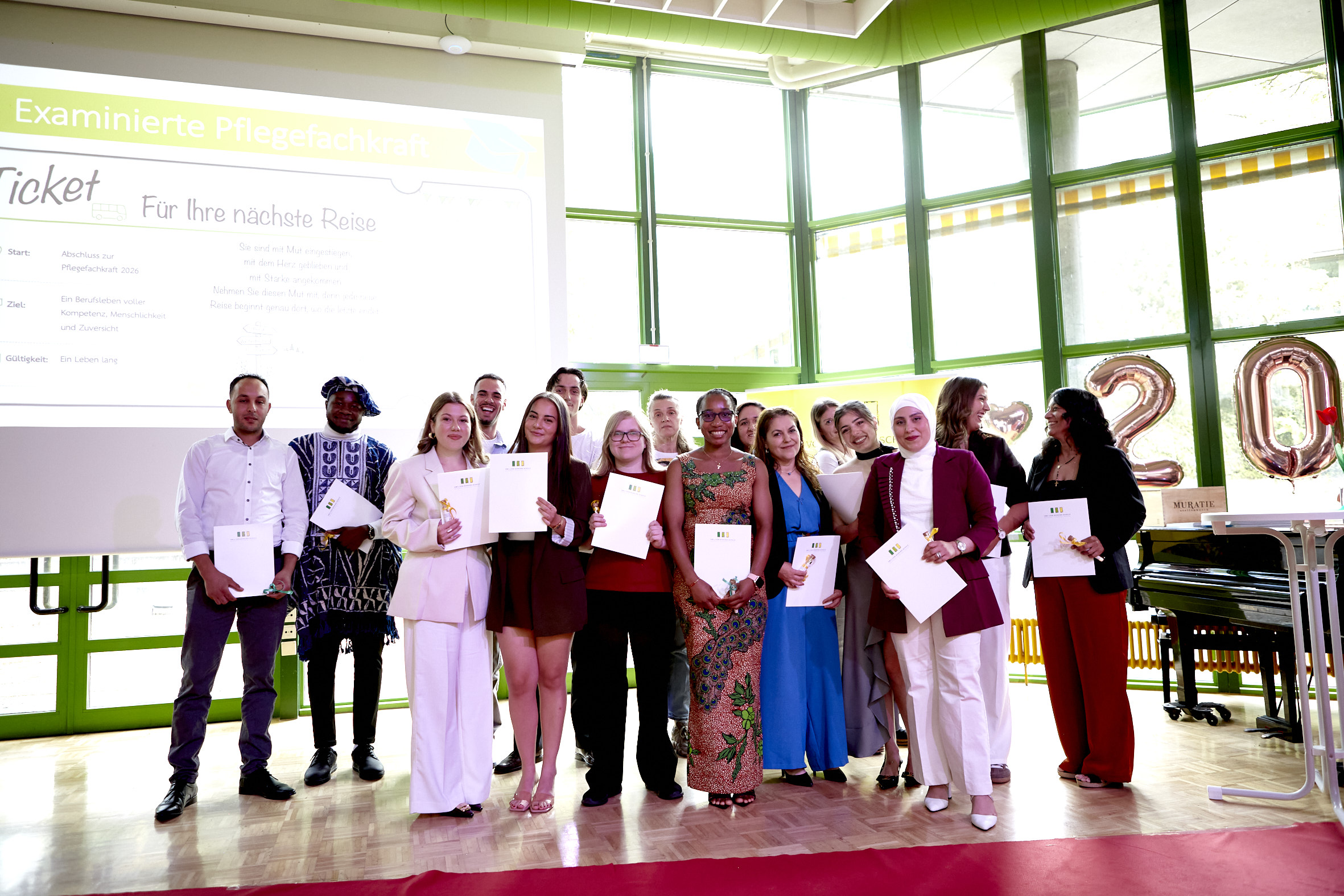 Absolventinnen und Absolventen der Pflegeausbildung stehen festlich gekleidet in einer Aula und präsentieren stolz ihre Urkunden. Im Hintergrund sind Projektionen und goldene Folienballons zu sehen.