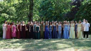 Eine große Gruppe von Absolventinnen und Absolventen steht nebeneinander im Grünen auf einer Wiese vor dichten Bäumen. Die Frauen tragen elegante, farbenfrohe Abendkleider und die Männer Anzüge bzw. Hemden. Alle blicken lächelnd in die Kamera.