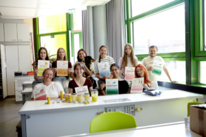 Eine Gruppe von neun lächelnden Personen, meist Schülerinnen und Schüler sowie zwei Lehrkräfte, posiert in einem hellen Klassenzimmer oder Fachraum für ein Foto. Sie stehen und sitzen hinter einem weißen Tisch, auf dem verschiedene Gläser mit gelbem Inhalt und Informationsblätter stehen. Viele halten Informationsblätter in die Kamera, auf denen Grafiken und Texte zu sehen sind. Im Hintergrund gibt es Schränke und große Fenster mit Blick nach draußen.