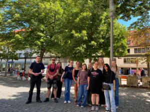 Eine Gruppe von elf Schülerinnen steht auf einem gepflasterten Platz vor einem historischen Gebäude mit großen Bäumen. Alle schauen in die Kamera und lächeln. Ein Schüler in einem Hard Rock Café T-Shirt steht im Vordergrund. Der Platz ist von Bäumen gesäumt, und im Hintergrund ist eine Bank und ein Straßenschild zu sehen. Das Licht ist hell und sonnig.