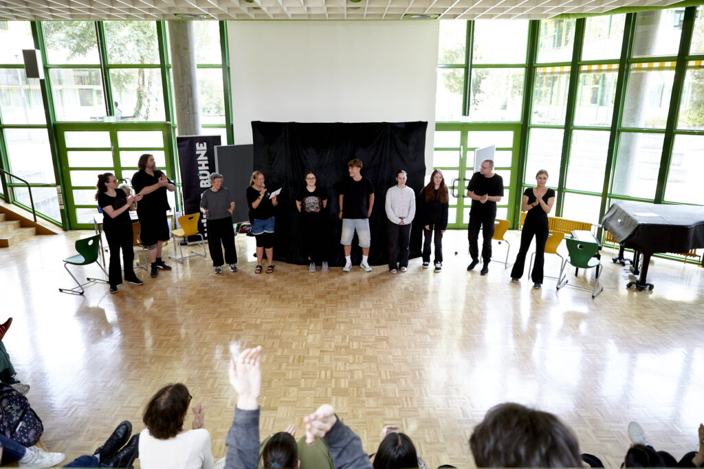 Eine Gruppe von Schauspielenden steht im Halbkreis in einer lichtdurchfluteten Schulaula vor schwarzem Bühnenhintergrund. Das Publikum im Vordergrund klatscht begeistert Beifall.